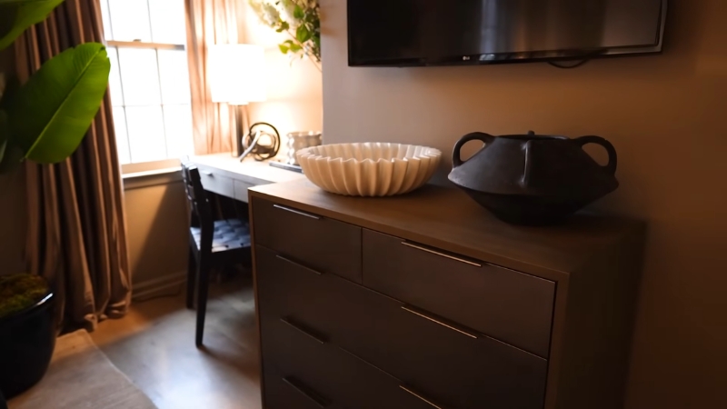 Komoda u maloj spavaćoj sobi sa toplim svetlom lampe pored prozora i radnim stolom u pozadini