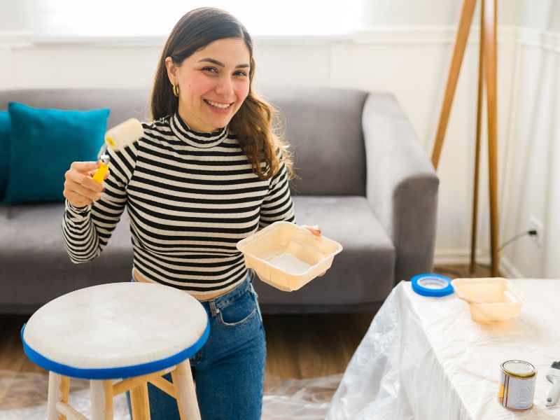 Srećna žena se smeši i uspostavlja kontakt očima dok farba stolicu i menja dekoracije prilikom preuređivanja nameštaja.