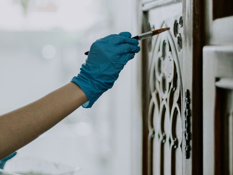 Jedna mlada belkinja, neprepoznatljiva, u plavim jednokratnim rukavicama, farba šablonska vrata starog ormara belom bojom uskom četkom, sedeći u sobi, krupni plan iz donjeg ugla.